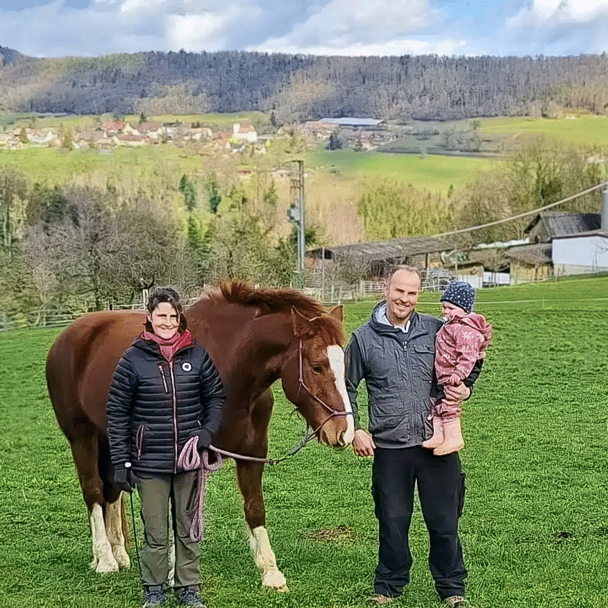 Auf dem Foto sind die Dacheigentümer:innen zu sehen: Rechts steht eine Frau mit dunklen Haaren. Sie hält ein braunes Pferd am Halfter. Rechts steht ein Mann, der ein Kleinkind auf dem Arm hält.
