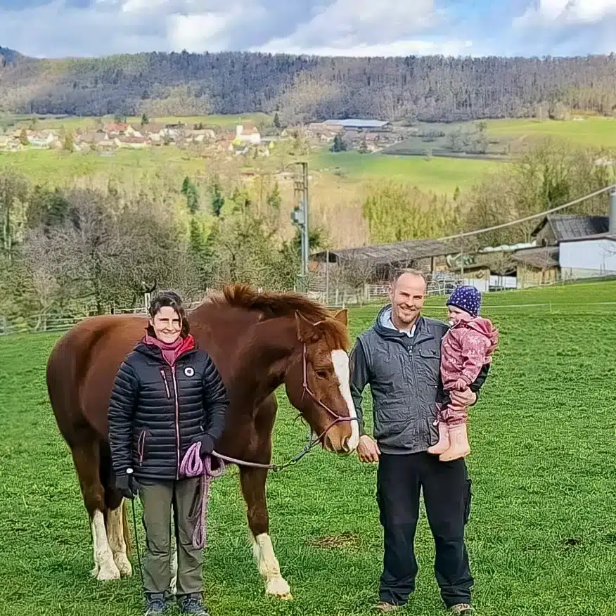 Auf dem Foto sind die Dacheigentümer:innen zu sehen: Rechts steht eine Frau mit dunklen Haaren. Sie hält ein braunes Pferd am Halfter. Rechts steht ein Mann, der ein Kleinkind auf dem Arm hält.
