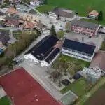 Aerial view of a small town: red multi-sport court in the foreground, row of houses with red-tiled roofs, parked cars, and stacks of lumber along a street.