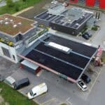Aerial view of a solar power installation on flat rooftops of an industrial complex, with rows of panels and nearby parked cars and vans.
