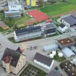 Aerial view of a town block with a red athletics track, green field, roads, and various white and gray buildings.