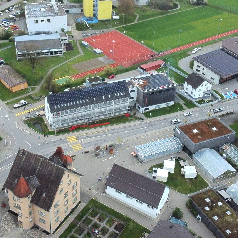 Aerial view of a town block with a red athletics track, green field, roads, and various white and gray buildings.