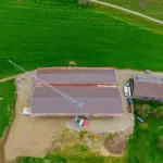 Aerial view of a construction site with a tall crane beside two long red-roofed buildings, dirt ground, and surrounding green fields.