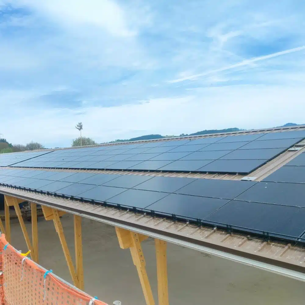 Rows of dark solar panels mounted on a sloped roof under construction, with wooden supports and an orange safety fence in the foreground.