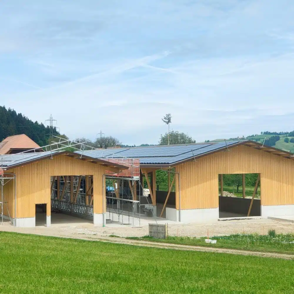 Two wooden buildings under construction with scaffolding and solar panels on the roof, set in a green field under a blue sky.