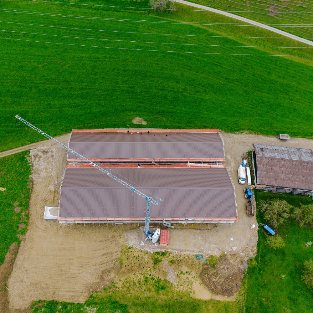 Aerial view of a construction site with a tall crane beside two long red-roofed buildings, dirt ground, and surrounding green fields.