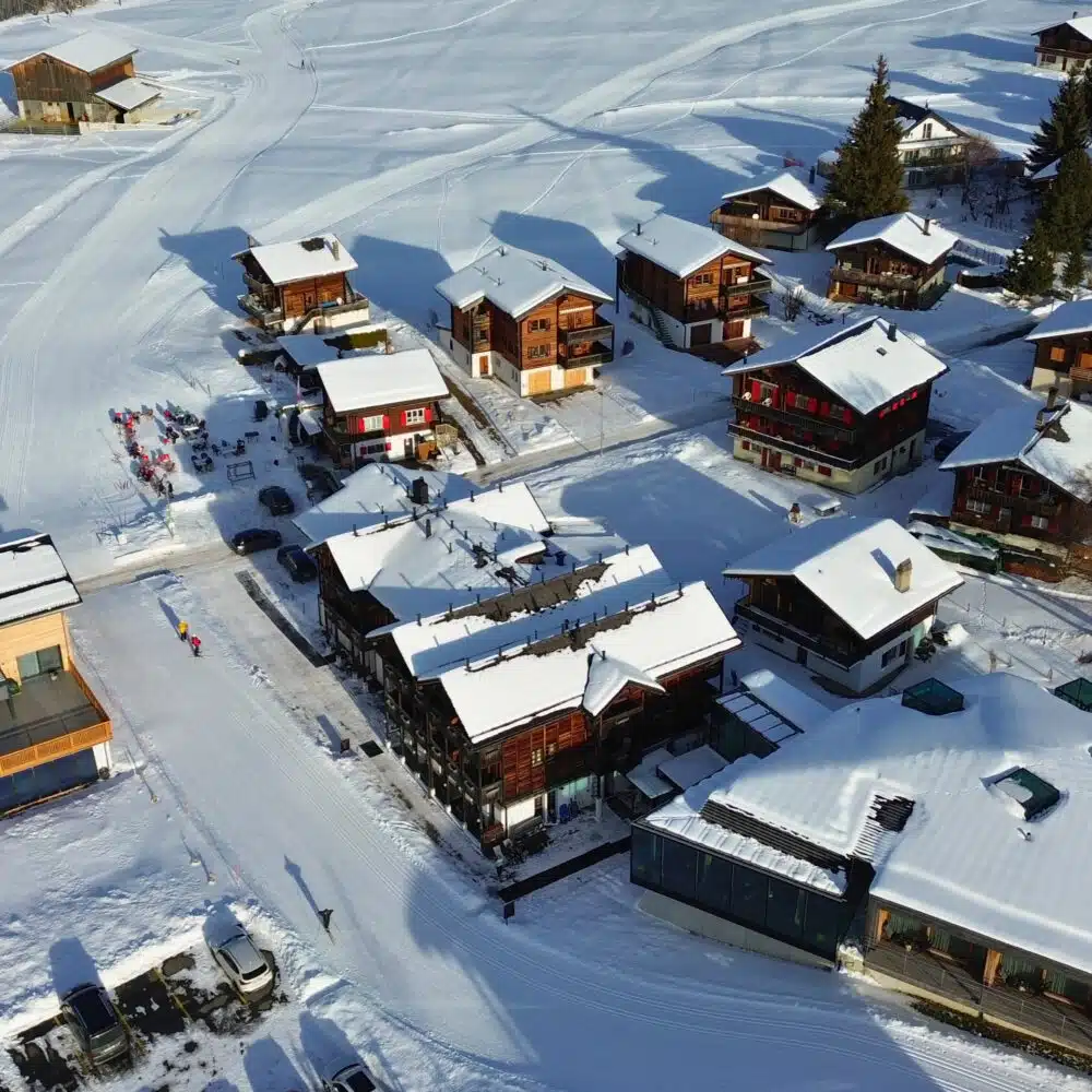 Aerial view of a snow-covered mountain village with wooden chalets, ski tracks across the slopes, and parked cars below.
