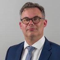 Martin-Hermann-Schwerzenbach Professional headshot of a man in a navy suit, white shirt, blue tie, wearing glasses against a gray background.