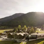 Sunlit alpine village with wooden chalets nestled in a green valley and forested hills beyond.