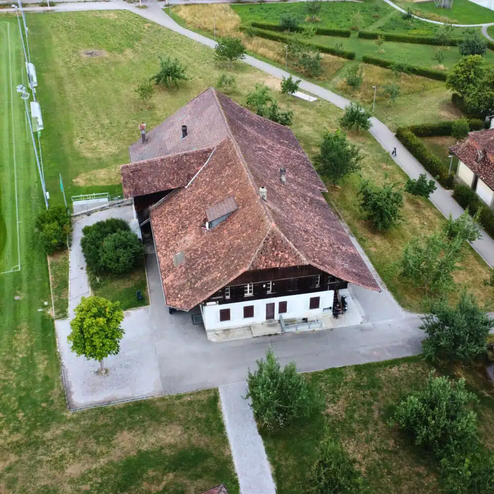 Vue aérienne d'un bâtiment blanc au toit de tuiles rouges, situé dans un parc verdoyant, avec des allées sinueuses et des arbres tout autour.