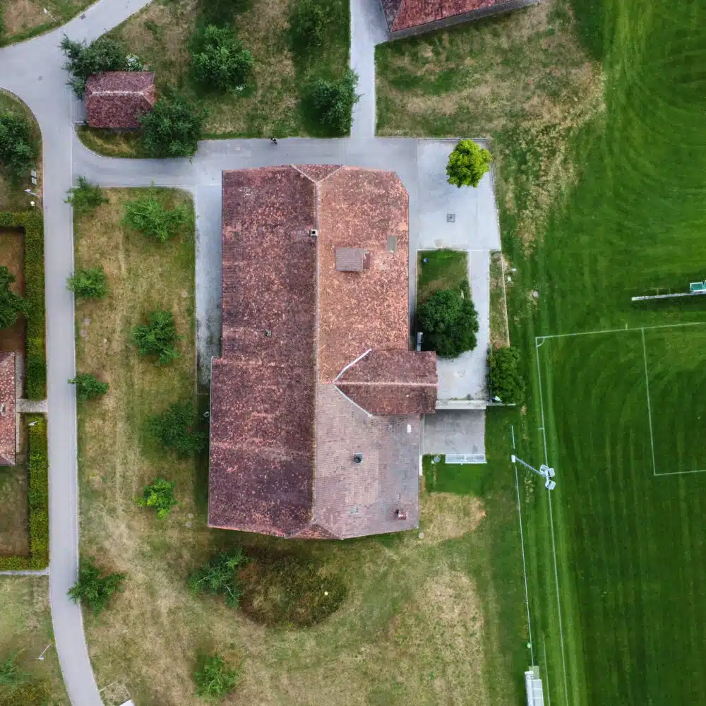Aerial view of a residential area with red-tiled roofs, curved roads, and surrounding green fields.