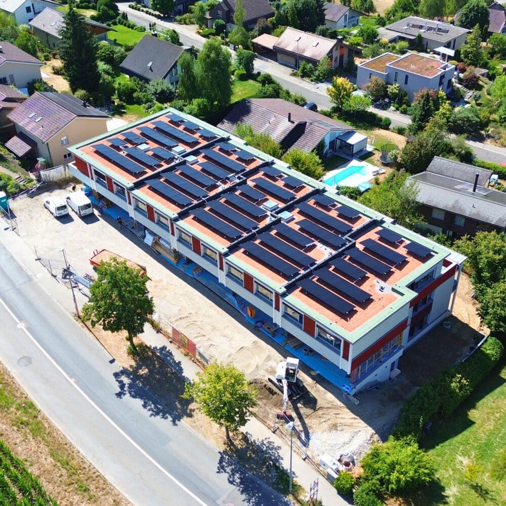 Aerial view of a residential neighborhood showing a large building with numerous solar panels on its roof under construction.