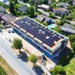 Aerial view of a residential neighborhood showing a large building with numerous solar panels on its roof under construction.