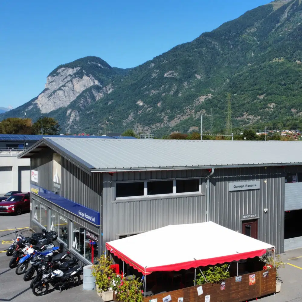 Bâtiment métallique gris devant lequel est garée une rangée de motos, avec au premier plan une tente blanche et rouge, sur fond de montagnes verdoyantes sous un ciel bleu limpide