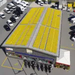 Un grand bâtiment doté d'un toit recouvert de panneaux solaires jaunes, avec un parking attenant où sont garées de nombreuses voitures et motos, ainsi que des places réservées aux personnes à mobilité réduite clairement signalées au premier plan.
